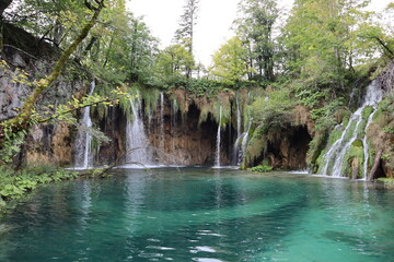 Scenic view of a lake overgrown with green trees with emerald turquoise clear water, and a waterfall with powerful jets of water, autumn at Plitvice Lakes National Park 
