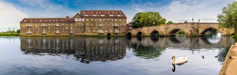 The mill and old bridge at Riverside, Godmanchester reflected in the calm waters of the River Great Ouse as a swan serenely glides past in springtime © Nicola