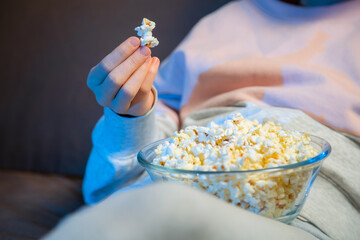 Close up of hand waking popcorn from a bowl while watching TV. Person sitting in comfortable couch and watching home cinema in the dark.