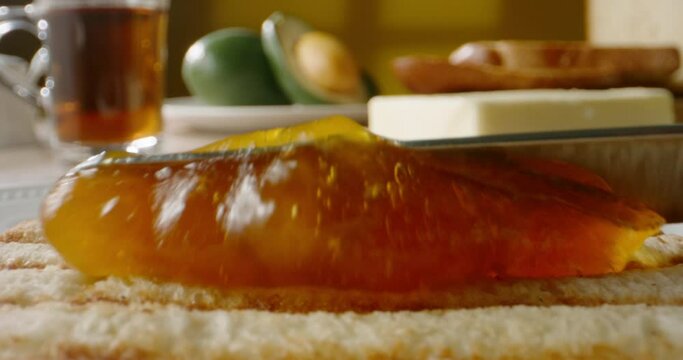 Preparing a sweet tasty sandwich for breakfast. Knife spreading apricot or peach jam on piece of fresh toast, close up 4k footage.