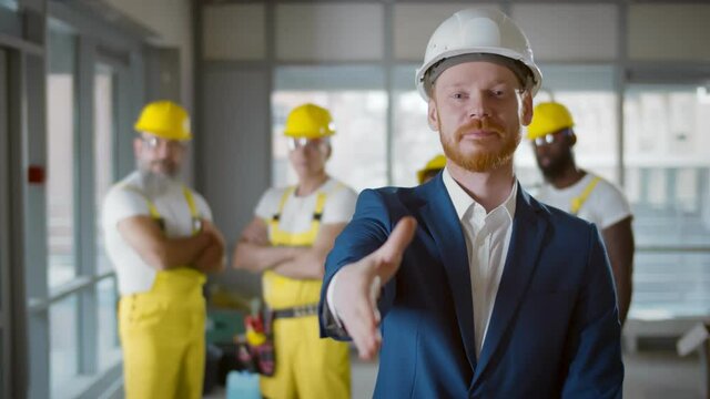 Contractor in hardhat stretching hand for handshake at camera with builders on background