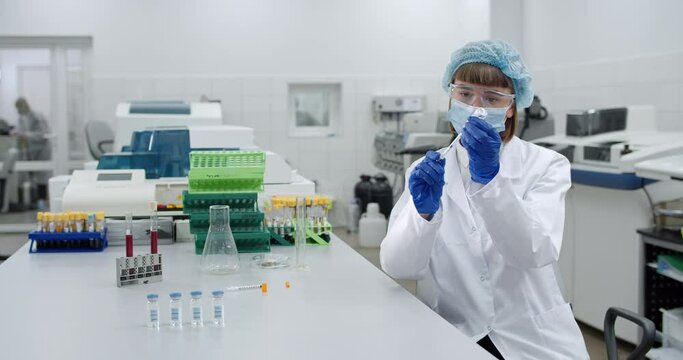 Crop view of female researcher in bouffant capfilling syringe with liquid from glass bottle. Woman in protective medical mask and white professional gown working in laboratory.