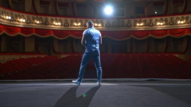 Man in front of empty theatre hall with red velvet chairs. Actor is standing on stage and talking with gestures before the performance.