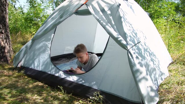 Young white kid playing online computer games using his mobile smartphone while laying inside of blue camping tent in summer green forest