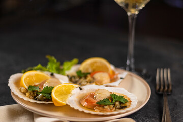 Scallops with lemon on black background with napkin and fork. Baked scallops with caviar in plate against background of blurred glasses of wine. Soft focus