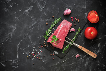 Raw beef fillet on a fork with spices, herbs and tomato. Black stone background. Fresh beef steak. Top view, flat lay.