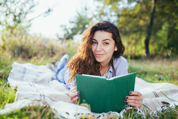 Obraz premium Beautiful young woman with curly black hair, resting lying on a mat in the park on the grass, and holding a book reads