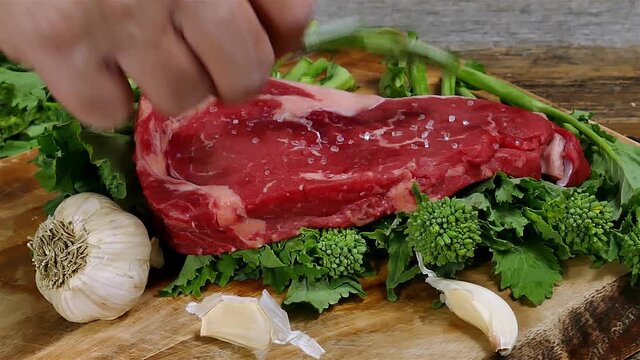 Hand is adding uncooked boneless club steak and rosemary on cutting board on babe broccoli and garlic around him