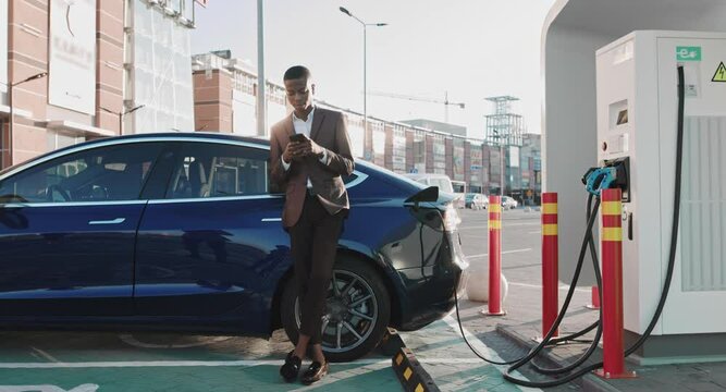 African American Businessman Using Smartphone And Leans On White Luxurious Electric Car That Is Charging. Plug Charging An Electrical Car. New Technologies At City Stops In Town Center