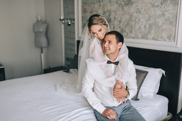 Fototapeta premium A beautiful, sexy bride in white lingerie hugs a stylish groom in a white shirt and black bow tie behind his back, sitting on a bed in a hotel room. Wedding portrait of newlyweds in love.
