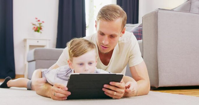 Smiling young American family lying on carpet with little kid watch cartoons on tablet, happy caucasian parent have fun relax with boy child on divan enjoy video on pad spending time at home together.