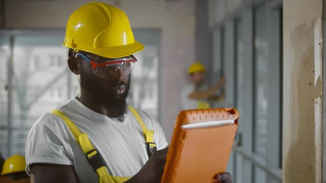 Afro-american builder in safety glasses and hardhat using tablet computer at construction site