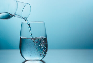 Fresh water pouring from the glass jug into a glass on the blue background with copy space