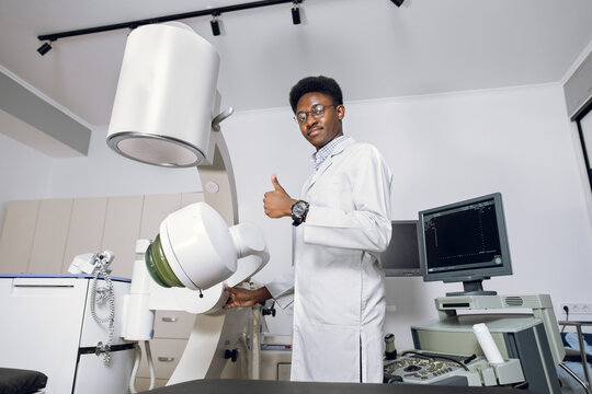 Portrait of handsome young professional African man doctor standing near modern ultrasound and lithotripter machines and showing thumb up. Safety non-invasive stone treatment, shock wave lithotripsy