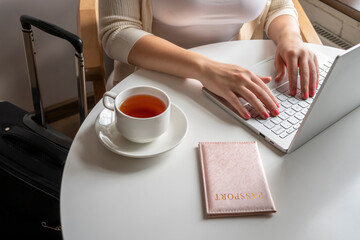 Woman tourist with suitcase having breakfast with cup of coffee putting passport on the table sitting near window in cafe in airport. Travel concept