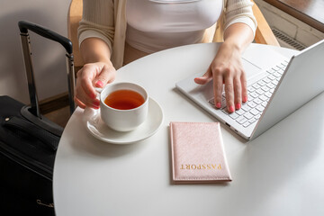 Woman tourist with suitcase having breakfast with cup of coffee putting passport on the table sitting near window in cafe in airport. Travel concept
