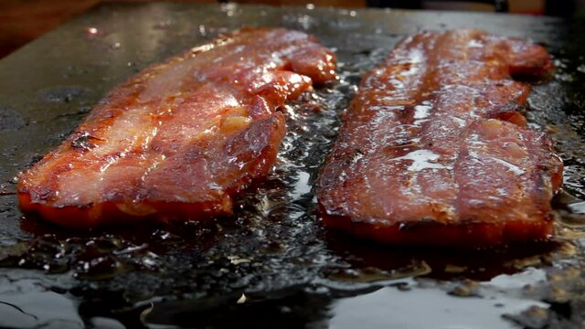 Close-up of bacon strips frying and sizzling on the flat grill surface outdoors