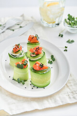Holiday vegetable appetizers. Cucumbers rolls with soft cheese, pieces of salted salmon, microgreens and black sesame served on a white plate. Selective focus.