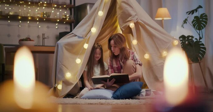 Mother and daughter are sitting in teepee tent reading stories