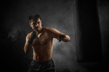Fototapeta na wymiar sporty shirtless boxer in black boxing wraps punching in boxing bag on dark background with smoke