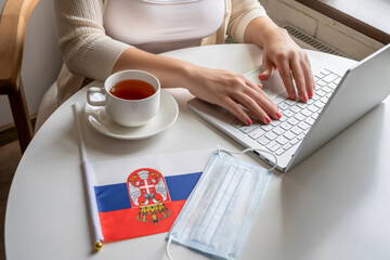 Woman tourist having breakfast with cup of coffee working on laptop.  Flag of Serbia, medical protective face mask on table of cafe. Protection from bacteria and virus in a public place. Concept.