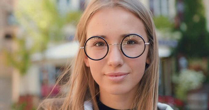 Close up of adorable cheerful happy young light-haired woman which putting on stylish glasses while posing on camera with sincerely smile outdoors