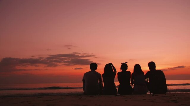 Back view silhouettes, group of young friends sitting and relaxing together on the beach at sunset.