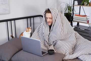 Young attractive caucasian man sitting on a bed wrapped with blanked at home working using laptop...