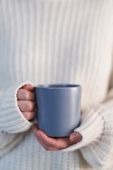 Obraz na płótnie Canvas Woman hands with a gray mug of hot drink. Beautiful girl in white sweater holding cup of tea or coffee in morning sunlight. Empty mug for your design. Concept o winter comfort, morning time