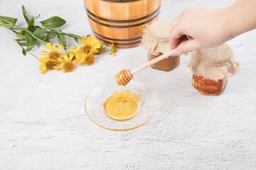 A woman's hand holds a wooden spoon over a saucer of honey, a ceramic barrel and jars of honey on a light background. Copy space