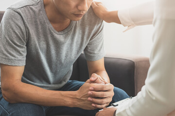 Young asian man, male suffers from a mental who needs to therapy with a psychologist while sitting on couch to consult, psychiatrist has encouragement the patient by touching to make his feel relaxed.