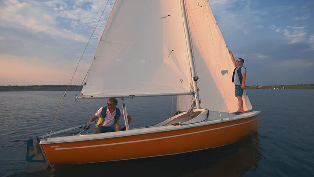 Orange sailing boat on a lake or river, windless weather, calm water without waves. Two sailors man operate the boat set sail and running rigging, enjoy vacation. Sailing yacht traveling on water