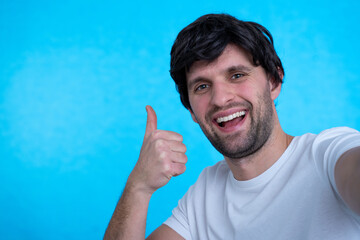 Naklejka premium Photo of handsome man in casual t-shirt and bristle on face smiling on camera with thumb up while taking selfie isolated over blue background