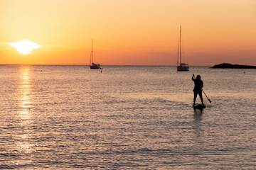 Man on stand up paddle board at sunset in calm waters with a sailboat