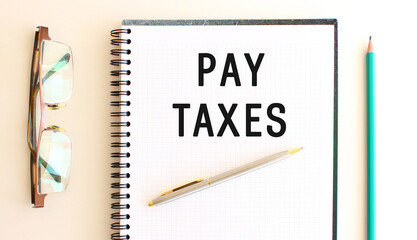 Notepad with text PAY TAXES on a white background, near glasses and pencil.