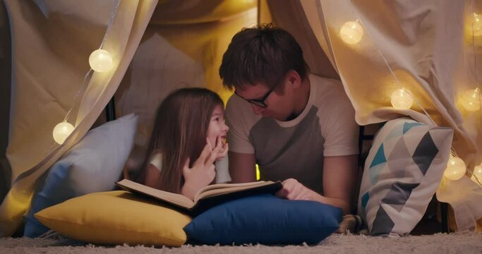 Happy father and daughter lying on floor in teepee and reading book together at home