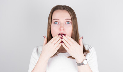 Young woman in a white T-shirt stands against a gray wall and covers her mouth with her hands in great surprise.