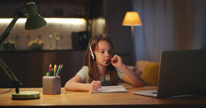 Little girl learning online with headphones and laptop sitting at desk at home doing homework