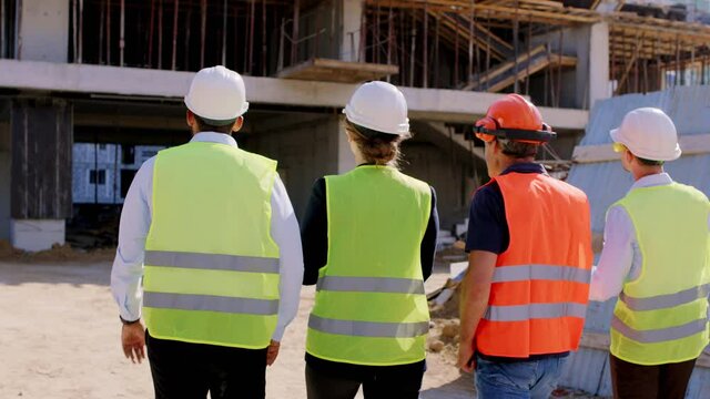 Taking video form the back of a group of diverse young specialists and foreman with ration checking the building construction while walking through the site they wearing protective uniform