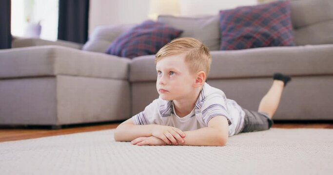 Cute little boy with blonde hair is lying on the floor staring at his favorite cartoon on the TV.
