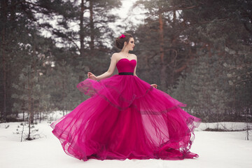 A beautiful girl in a cyclamen color ball gown on the background of a snow-covered winter forest.