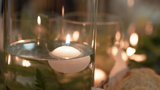Floating white wax candles are in a transparent glass vase with water, a flame is burning on the wick