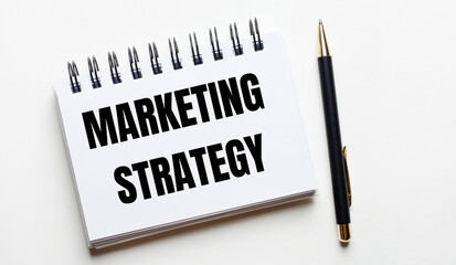 On a light background, a white notebook with the words MARKETING STRATEGY and a pen.