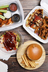 Top view of table fool of food in plastic containers. Food delivery. Chicken salad, chicken wings with sous, burger on french fries on a wooden table. Business lunch.