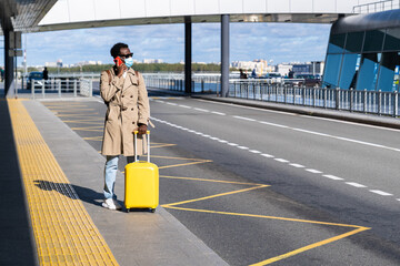 Afro-American millennial man with yellow suitcase stands in airport terminal, using phone, calling...