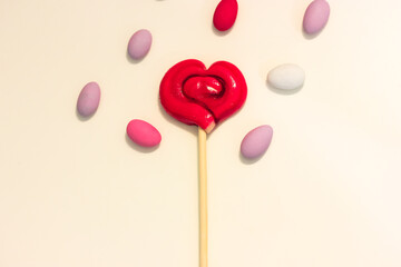Lollipop on a stick in the shape of a red heart on a white table surrounded by colorful candy, layout. A sweet treat for Valentine's Day 