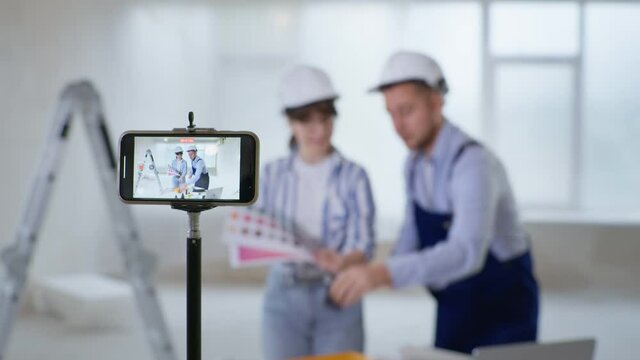 builders blog, smiling man and woman talk about interior design and renovation and show wall painting tool and color half on smartphone camera while recording video in construction site