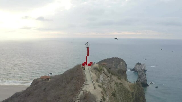 Aerial view of a red and white colored lighthouse with many vulture flying around the light house, heading towards the open ocean