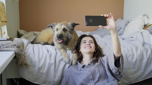 Happy young lady in grey shirt with smartphone makes selfie with large dog leaning on bed near window. Concept owner spend time with pet at home