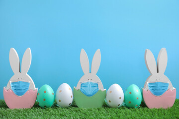 COVID-19 pandemic. Easter bunny figures in protective masks and dyed eggs on green grass against light blue background